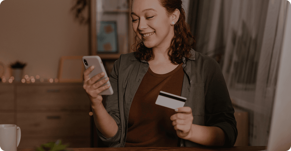 O femeie care ține un card de credit și un telefon mobil, zâmbind.